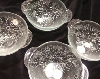 May include: Four clear glass bowls with handles, each featuring a poinsettia design. The bowls are round with a slightly flared rim and are set against a dark background. The poinsettia design is etched into the glass.