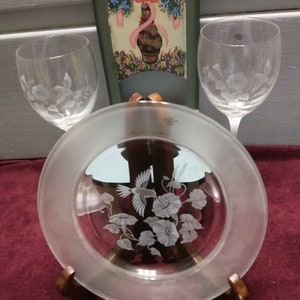 May include: A set of three clear glass etched pieces: a plate with a hummingbird and flowers, and two wine glasses with floral designs. The plate is on a wooden stand.