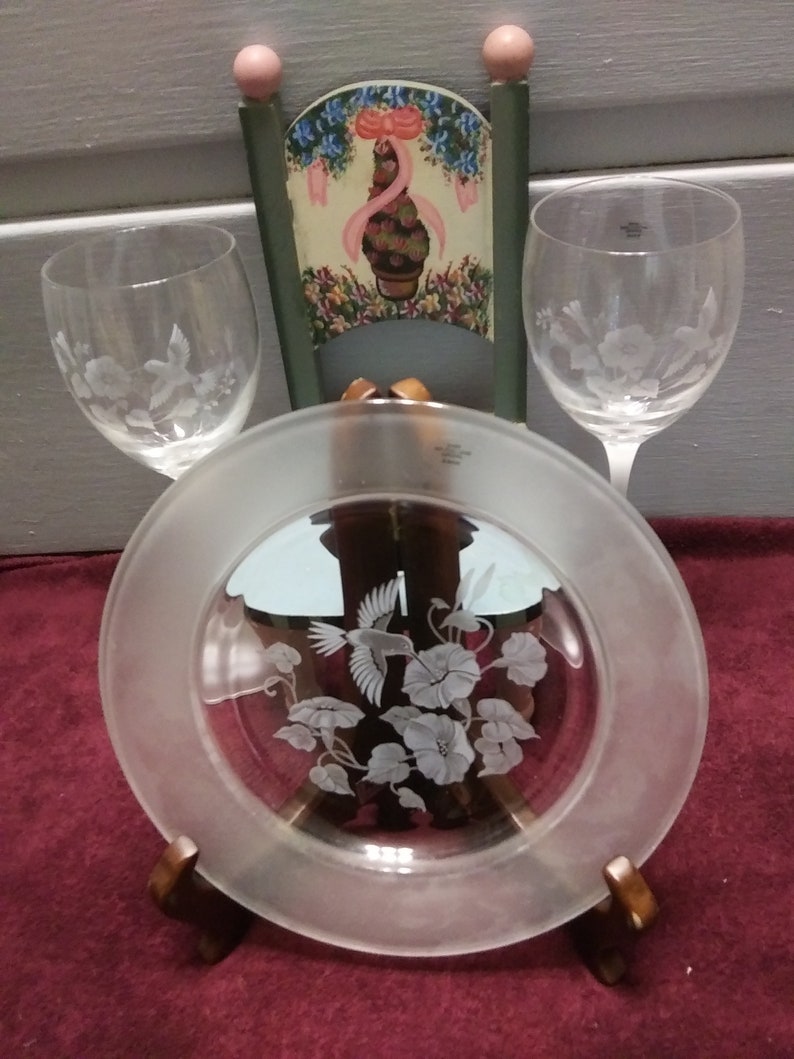 May include: A set of three clear glass etched pieces: a plate with a hummingbird and flowers, and two wine glasses with floral designs. The plate is on a wooden stand.