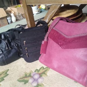 May include: Three handbags are displayed: a black bucket bag with silver hardware and drawstring, a black multi-zippered crossbody bag, and a magenta shoulder bag featuring a V-shaped fringe detail, braided trim, and a matching side tassel.