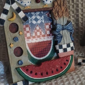 May include: A wooden wall decor featuring a whimsical farmhouse theme. The decor includes a birdhouse, a watermelon, wheat stalks, and a blue and white checkered pattern.