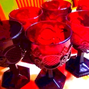 May include: Set of six ruby red goblets. Each glass features a decorative pattern on the bowl and a hexagonal base. The glasses are arranged on a white surface, with a colorful background.