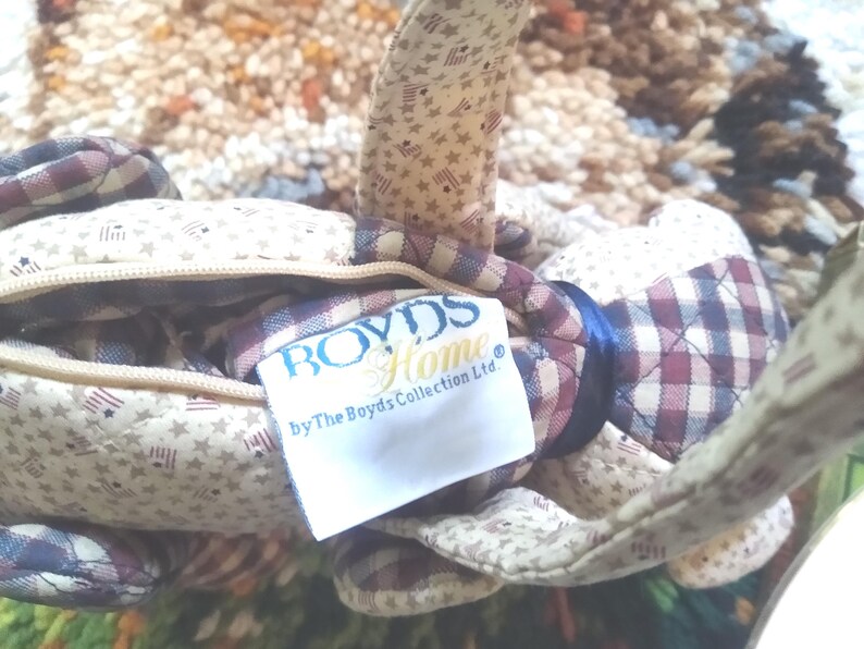 Puede incluir: Bolso de tela acolchada con cierre de cremallera. El bolso presenta un estampado de estrellas y rayas en beige, rojo y azul. La etiqueta dice "BOYDS Home by The Boyds Collection Ltd."