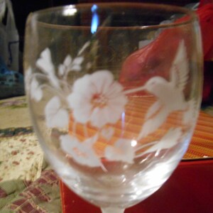May include: A clear glass wine glass with an etched design of a hummingbird and flowers. The glass has a frosted finish and is sitting on a red surface.