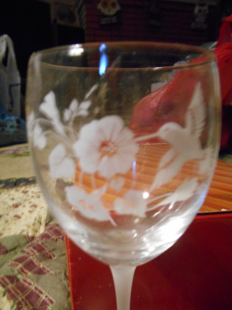 May include: A clear glass wine glass with an etched design of a hummingbird and flowers. The glass has a frosted finish and is sitting on a red surface.