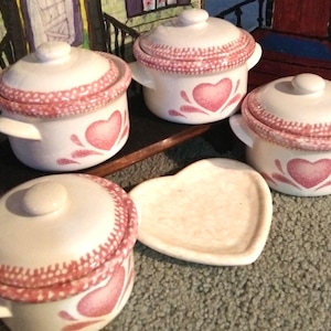 May include: A collection of white ceramic mini casserole dishes with lids, adorned with a pink heart design and a decorative pink trim. A heart-shaped serving dish is also present.