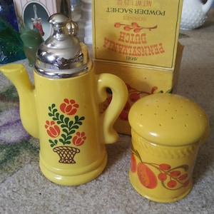 May include: Two yellow ceramic perfume bottles with floral designs. The larger bottle is shaped like a teapot with a silver lid. The smaller bottle has a round shape with a perforated lid. The bottles are labeled "Avon Pennsylvania Dutch Sonnet Powder Sachet Net Wt 1.25 oz."