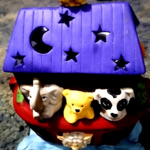 May include: A colorful ceramic Noah's Ark figurine. The ark has a red base, a purple roof with star cutouts and a crescent moon. Animals include an elephant, bear, cow, and sheep. Two yellow birds sit on top.