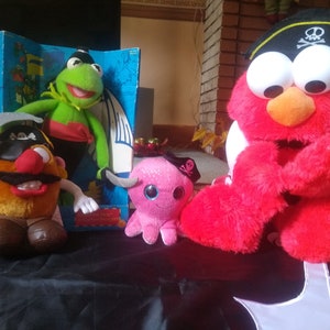 May include: Four plush toys dressed as pirates. A green frog, a red monster, a brown potato head, and a pink octopus. The toys are all wearing pirate hats and have swords.