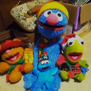 May include: A group of plush Sesame Street characters, including Grover, Cookie Monster, and Kermit the Frog, wearing winter hats and scarves. A yellow McDonald's Baby Fozzie Bear toy is also present. The characters are arranged on a light-colored surface.