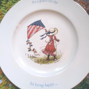 Puede incluir: Un plato decorativo blanco con borde dorado presenta a una ni&ntilde;a volando una cometa con un dise&ntilde;o de bandera estadounidense. El plato tiene el texto "It's a grand old day" y "for being happy".