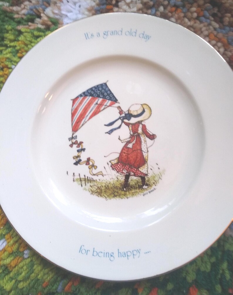 Puede incluir: Un plato decorativo blanco con borde dorado presenta a una ni&ntilde;a volando una cometa con un dise&ntilde;o de bandera estadounidense. El plato tiene el texto "It's a grand old day" y "for being happy".