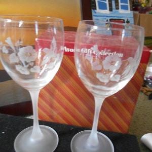 May include: Two clear glass wine glasses with frosted bases. The glasses are etched with a hummingbird and floral design. The glasses are sitting on a black surface in front of a red and orange striped box with the text "Avon Gift Collection".