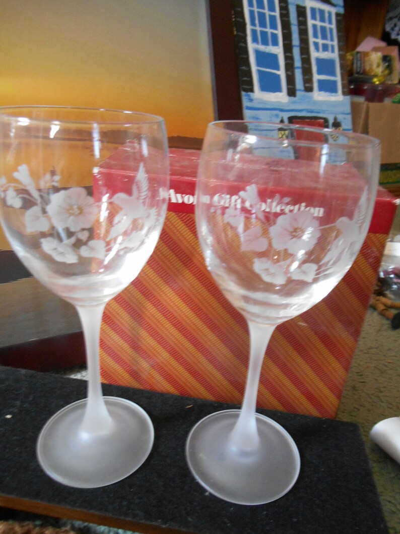 May include: Two clear glass wine glasses with frosted bases. The glasses are etched with a hummingbird and floral design. The glasses are sitting on a black surface in front of a red and orange striped box with the text "Avon Gift Collection".