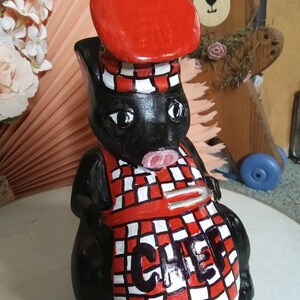May include: A black pig figurine wearing a red chef's hat and apron with a red and white checkered pattern. The apron has the word "CHEF" in black. The pig has white eyes and a pink snout. A wooden bear is in the background.