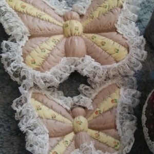 May include: Two butterfly-shaped quilted pot holders. The pot holders are mainly pink with yellow and floral accents. Each butterfly has a white lace trim around the edges. They are designed to protect surfaces from heat.