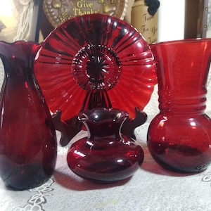 May include: Four pieces of ruby red glass, including a vase, a small bowl, a larger vase, and a decorative plate. The plate has a sunburst design.