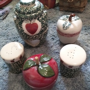 May include: Set of five ceramic apple-themed kitchen containers. The largest container is a green and white jar with a red heart and a cutout heart on the side. There are two smaller white salt and pepper shakers with green and white designs. The other two containers are shaped like apples, one is red with green leaves and the other is white with green leaves.