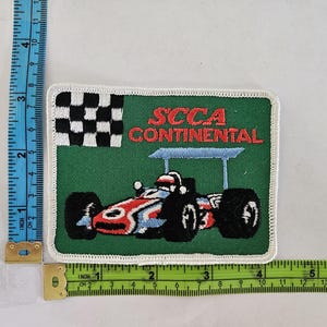 May include: Embroidered patch featuring a race car with the text "SCCA CONTINENTAL" and a checkered flag. The patch is rectangular, with a green background and white border. The race car is red, white, and blue.