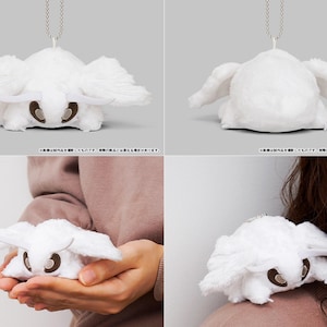 May include: White plush toy of a moth with a silver chain attached. The moth has large wings and a small, round body. The toy is shown in different positions, including hanging from the chain and being held by a person.