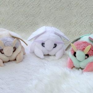 May include: Three plush toys shaped like moths. The first moth is cream colored with a light brown head and wings. The second moth is white with light purple wings. The third moth is pink with light green wings.