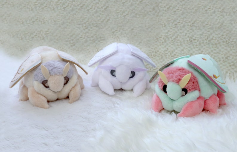 May include: Three plush toys shaped like moths. The first moth is cream colored with a light brown head and wings. The second moth is white with light purple wings. The third moth is pink with light green wings.