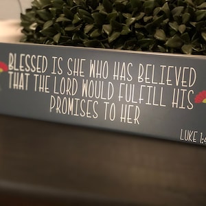 Könnte beinhalten: Ein dunkelblaues Holzschild mit weißem Text, der lautet "Blessed is she who has believed that the Lord would fulfill his promises to her" und "Luke 1:45". Das Schild hat rechts ein rotes Blumenmuster.