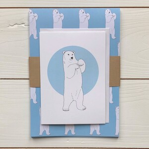 May include: A white card with a blue circle and a black and white illustration of a polar bear standing on its hind legs. The card is on top of a blue wrapping paper with a repeating pattern of polar bears.