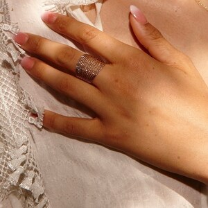 Op de afbeelding: Een goudkleurige ring met een geweven ontwerp wordt op een hand gedragen. De ring is gemaakt van meerdere dunne bandjes, waardoor een textuur ontstaat. De hand rust op een wit kledingstuk met kanten rand.