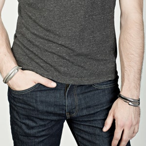 Pu&ograve; includere: Una persona indossa una maglietta grigio scuro e jeans blu scuro. La persona indossa due braccialetti d'argento. Un braccialetto &egrave; sul polso sinistro e l'altro sul polso destro.