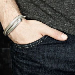 Pu&ograve; includere: Un bracciale argentato con un motivo a spina di pesce strutturato. Il bracciale &egrave; indossato su un polso, con la mano appoggiata nella tasca di un paio di jeans blu scuro. La persona indossa una maglietta grigia.
