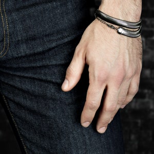 Pu&ograve; includere: Un braccialetto argentato con un design a forma di serpente &egrave; indossato al polso di una persona. Il braccialetto ha un aspetto a strati ed &egrave; collegato da una piccola catena. La persona indossa jeans di denim blu scuro.