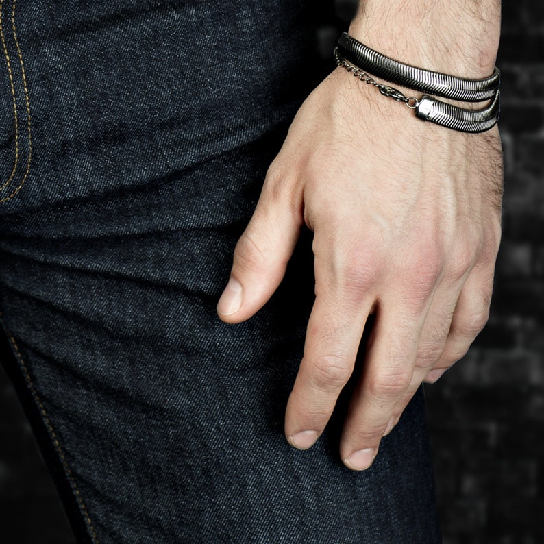 Pu&ograve; includere: Un braccialetto argentato con un design a forma di serpente &egrave; indossato al polso di una persona. Il braccialetto ha un aspetto a strati ed &egrave; collegato da una piccola catena. La persona indossa jeans di denim blu scuro.