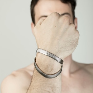 Pu&ograve; includere: Due braccialetti piatti color argento a catena a serpente sono indossati sul braccio di una persona. I braccialetti hanno un motivo testurizzato e ondulato. Lo sfondo &egrave; di colore grigio chiaro.