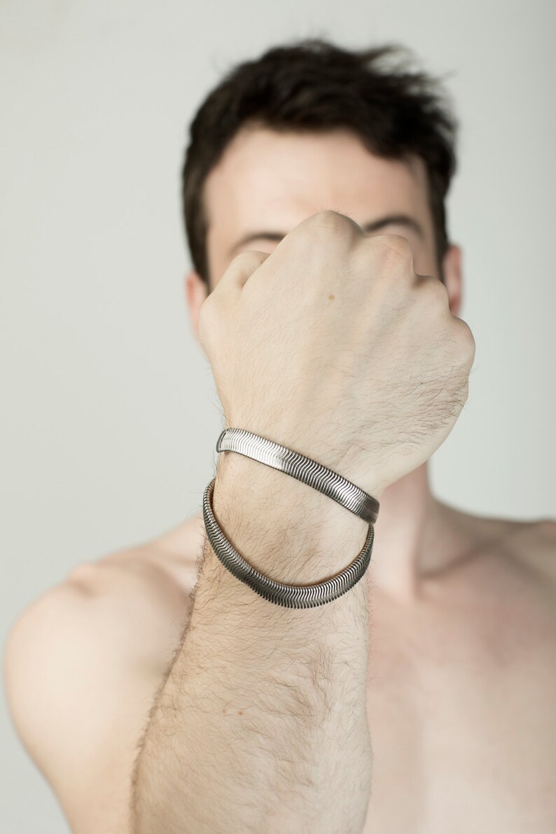 Pu&ograve; includere: Due braccialetti piatti color argento a catena a serpente sono indossati sul braccio di una persona. I braccialetti hanno un motivo testurizzato e ondulato. Lo sfondo &egrave; di colore grigio chiaro.