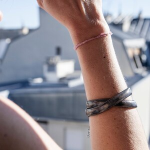 Pu&ograve; includere: Un braccialetto in metallo argentato con un design intrecciato &egrave; indossato su un braccio. &Egrave; visibile anche un delicato braccialetto rosa. Lo sfondo mostra tetti e un cielo soleggiato.