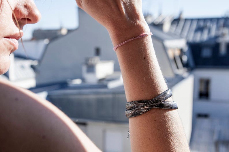 Pu&ograve; includere: Un braccialetto in metallo argentato con un design intrecciato &egrave; indossato su un braccio. &Egrave; visibile anche un delicato braccialetto rosa. Lo sfondo mostra tetti e un cielo soleggiato.