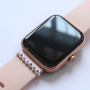 Może przedstawiać: Różowo-złoty smartwatch z czarną tarczą i różowym silikonowym paskiem. Srebrny pasek z fioletowymi serduszkami jest przymocowany do paska.