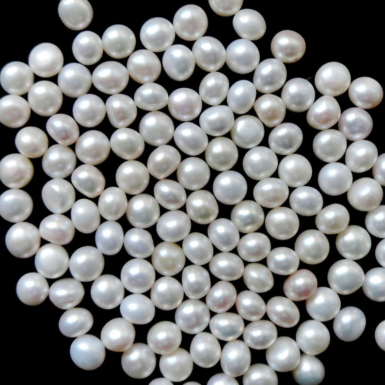 May include: A large group of white, round pearls scattered on a black background. The pearls are smooth and glossy, and they are all about the same size.
