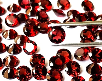 May include: A collection of round, red gemstones, likely garnets, scattered on a white surface. Some of the gemstones are being held by a pair of tweezers.
