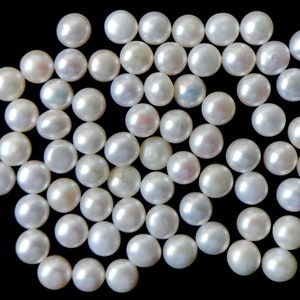 May include: A collection of white pearl beads scattered on a black background. The pearls are round and smooth, with a slight iridescent sheen.