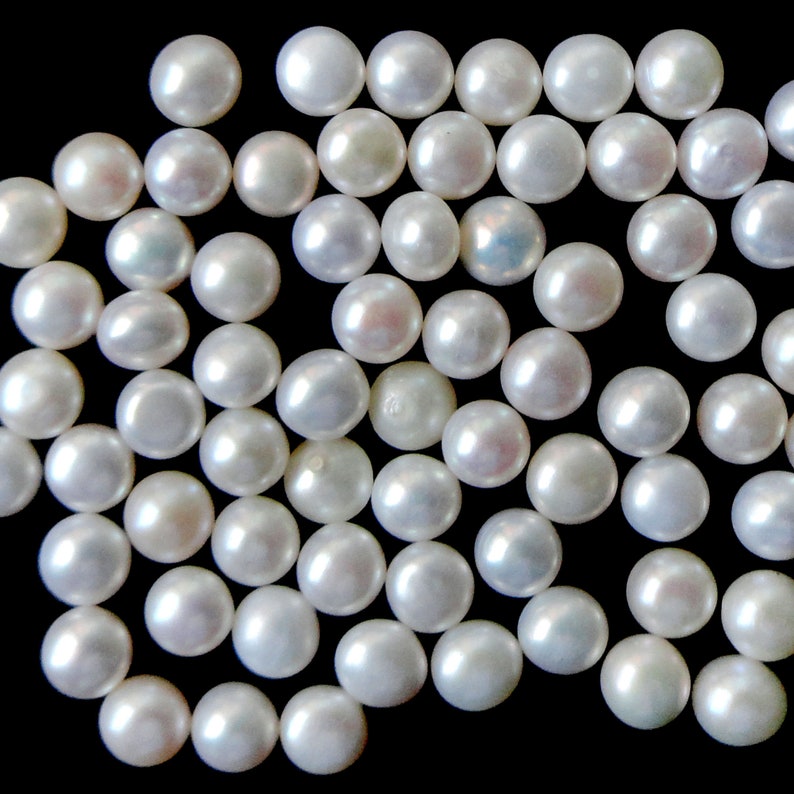 May include: A collection of white pearl beads scattered on a black background. The pearls are round and smooth, with a slight iridescent sheen.