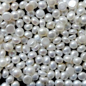 May include: A close-up image of a large quantity of white round pearl cabochons. The pearls are smooth and have a slight iridescent sheen.