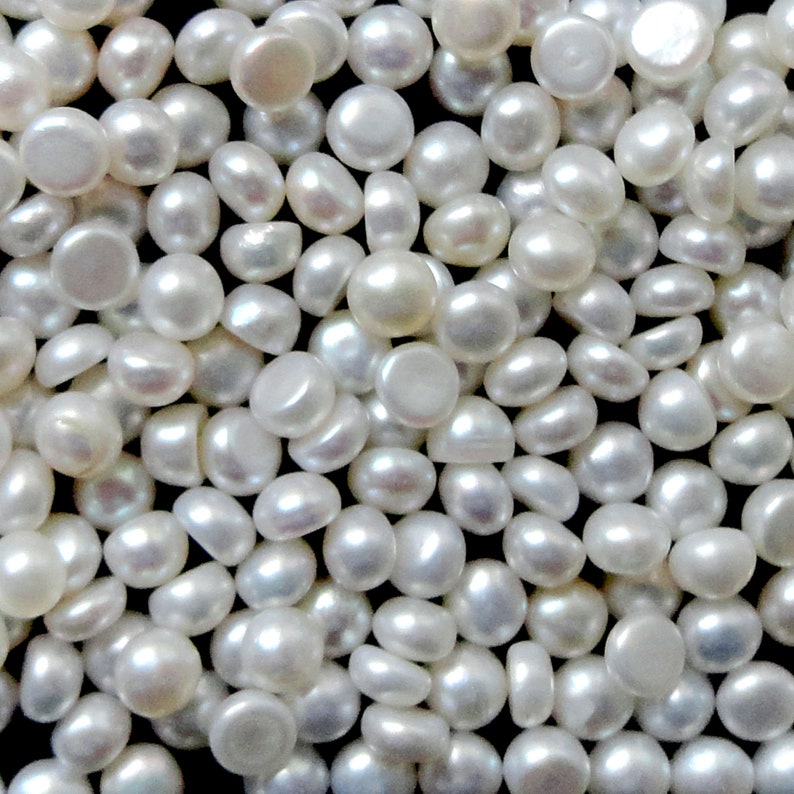 May include: A close-up image of a large quantity of white round pearl cabochons. The pearls are smooth and have a slight iridescent sheen.