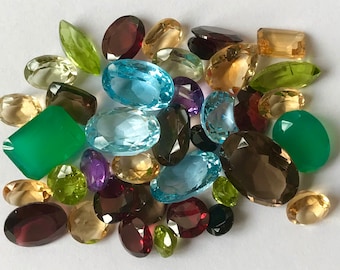 May include: A collection of various gemstones in shades of green, blue, yellow, purple, and brown. The gemstones are oval, rectangular, and faceted, with a variety of cuts and sizes. The gemstones are scattered on a white surface.