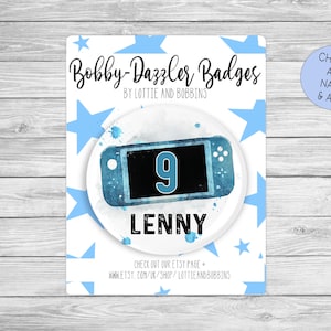 May include: A blue and white watercolor-style badge with a blue Nintendo Switch console and the number 9. The badge says "Bobby-Dazzler Badges" and "Lenny" in black text.
