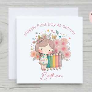 Puede incluir: Una tarjeta blanca con una ilustraci&oacute;n rosa y blanca de una ni&ntilde;a sosteniendo l&aacute;pices de colores. El texto "Happy First Day At School!" est&aacute; escrito en rosa encima de la ni&ntilde;a. El nombre de la ni&ntilde;a "Bethany" est&aacute; escrito en rosa debajo de la ni&ntilde;a.