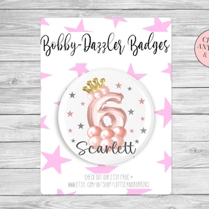 May include: A pink and gold birthday badge featuring a crown and the number 6. The name "Scarlett" is written below the number. The badge is surrounded by pink and grey stars.