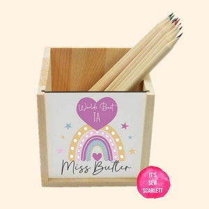May include: A wooden pencil holder with a rainbow and heart design. The holder has the text "World's Best TA" and "Miss Butler" on it.