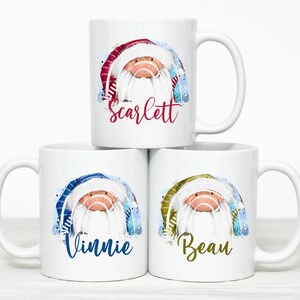May include: Three white ceramic mugs with a winter holiday theme. Each mug features a cartoon Santa Claus with a blue, red, or gold hat and a white beard. The mugs are personalized with the names "Scarlett", "Vinnie", and "Beau".